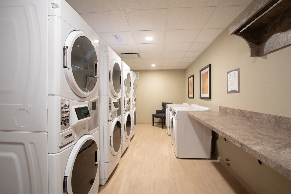A practical interior scene featuring modern laundry machines, a folding counter, and a comfortable seating area. The space is clean, communal, and designed for ease, supporting guests’ needs for fresh clothes during extended stays.