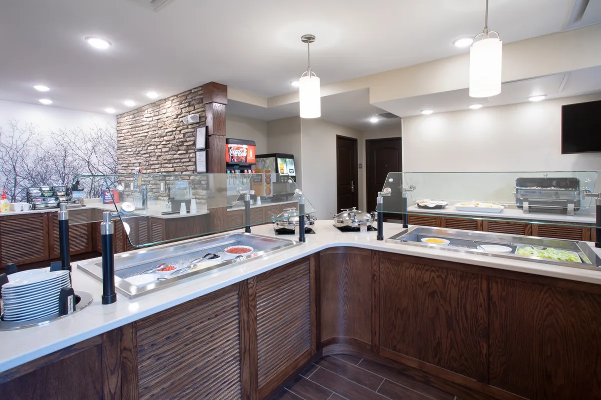 A bright interior scene featuring a buffet counter lined with various breakfast food selections, a beverage station, and a decorative stone wall in the background. Guests are seen helping themselves to the self-service breakfast and socializing over their meals in a casual, comfortable setting.