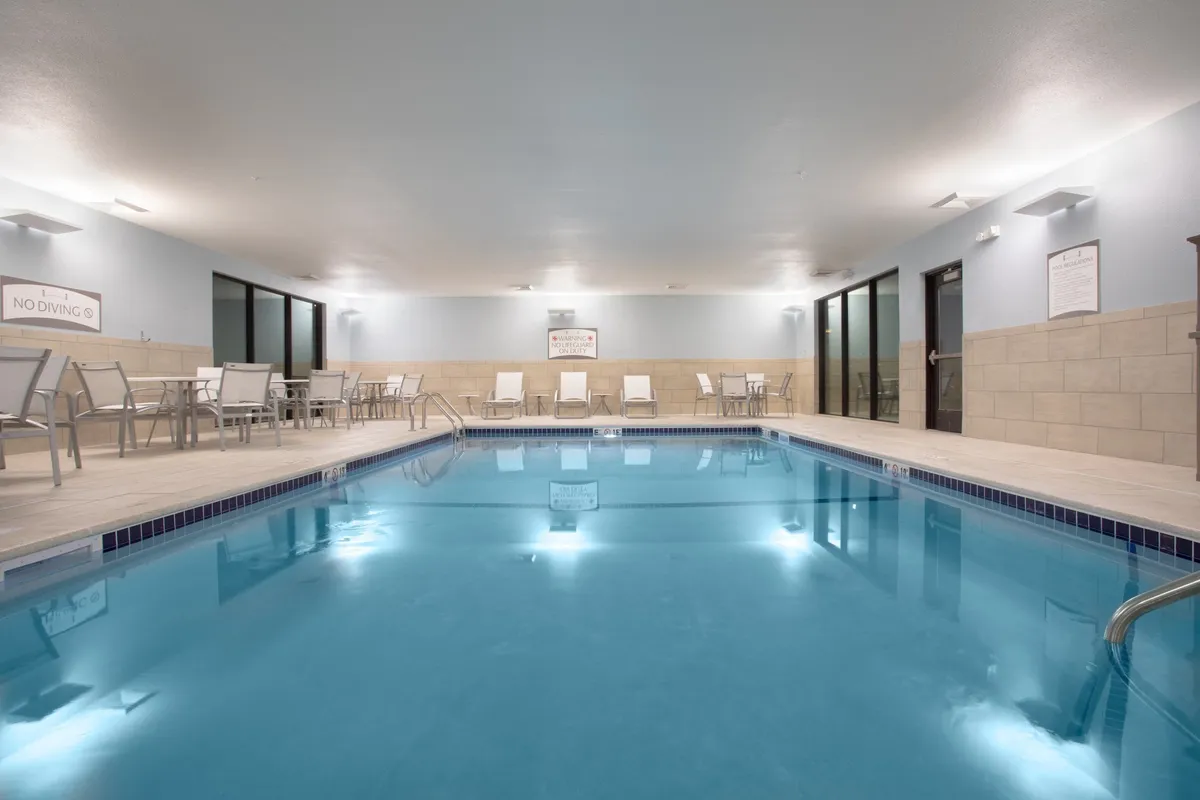 A serene indoor swimming pool area bordered by lounge chairs and surrounded by bright, inviting lighting. The space is designed for year-round enjoyment—ideal for swimming, relaxing by the water, or socializing with others.