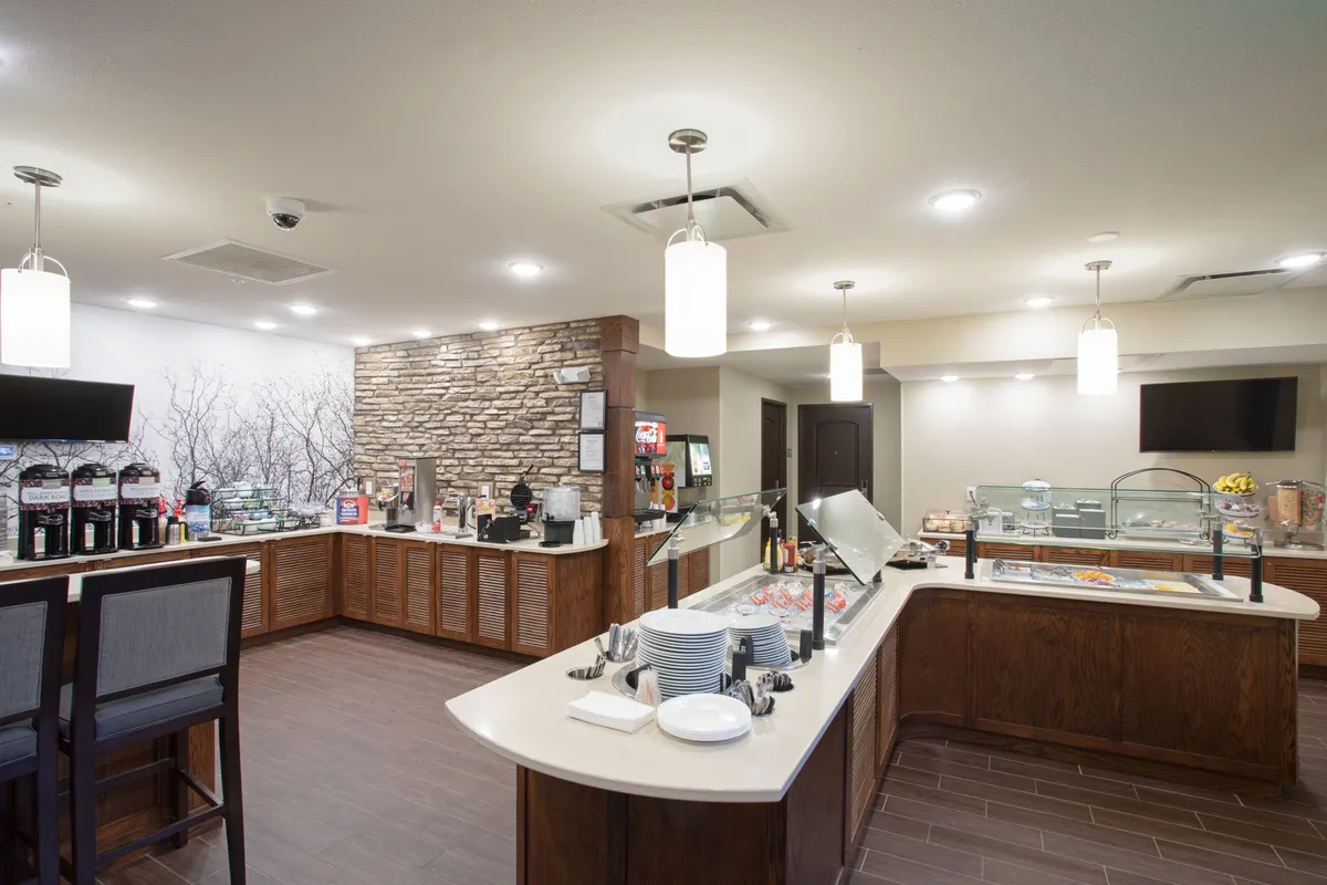 A warmly lit dining area showcasing a stone accent wall, a well-stocked breakfast bar, a coffee station, and an inviting food display. Guests gather at the breakfast bar, enjoying their meals and friendly conversation in a cozy, social environment.