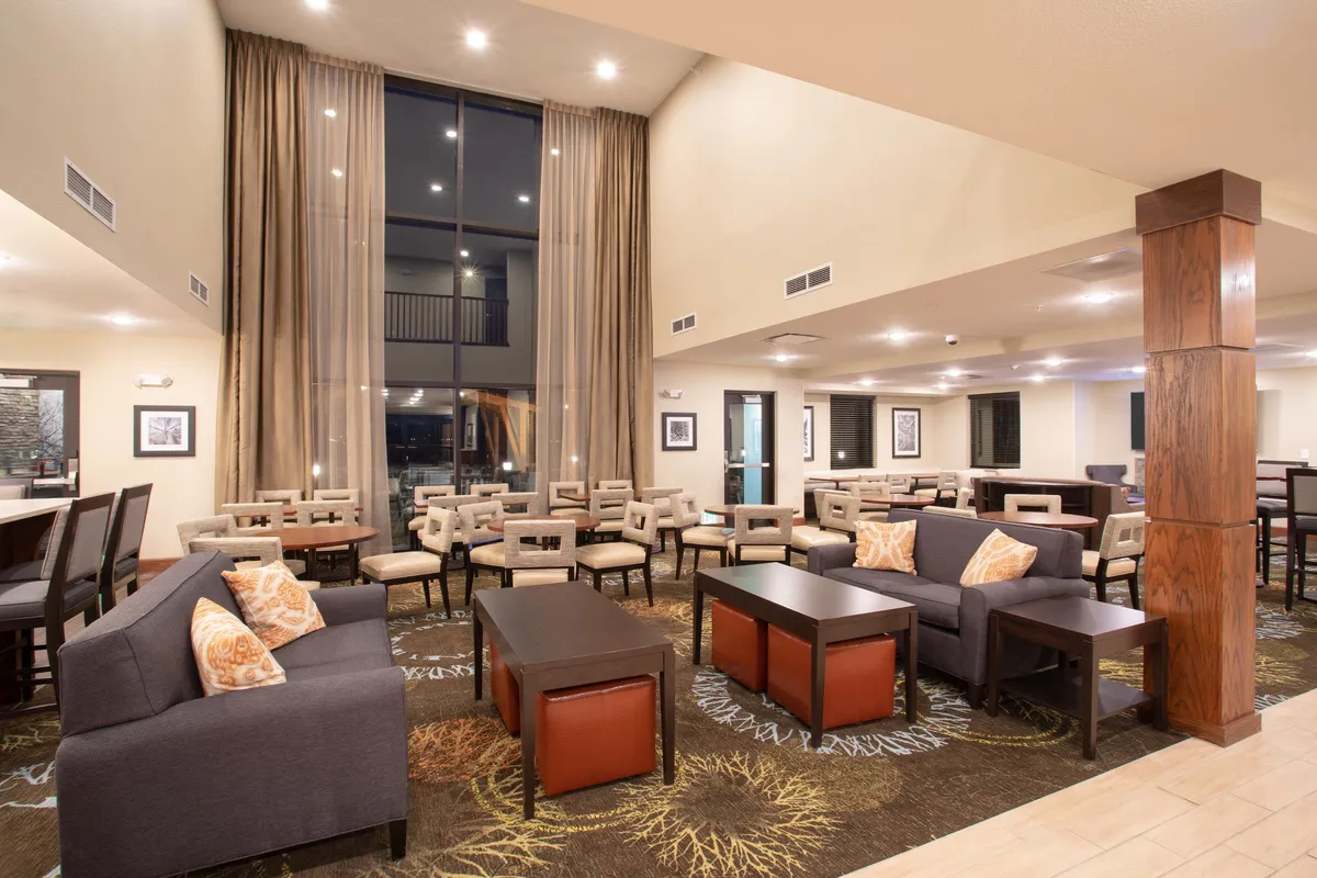 A cozy interior scene featuring a comfortable seating arrangement centered around a large window with curtains, accented by warm wooden pillars. The space is arranged to encourage socializing, reading, or enjoying coffee, with plush chairs and a relaxed atmosphere.