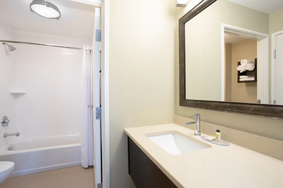 A modern bathroom with a sleek sink, wide mirror, and a bathtub. Fluffy towels are neatly arranged, and the space exudes tranquility, suggesting a perfect spot for a refreshing morning routine or a relaxing soak after a long day.
