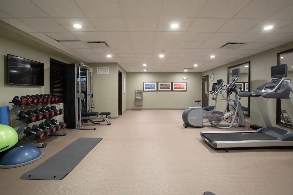 A well-equipped hotel fitness center featuring a dumbbell rack, treadmills, and elliptical machines. The space is bright and energetic, with workout equipment arranged for both cardio and strength training. Artwork on the walls adds a welcoming touch, and the open layout suggests an inviting place for guests to stay active during their travels.